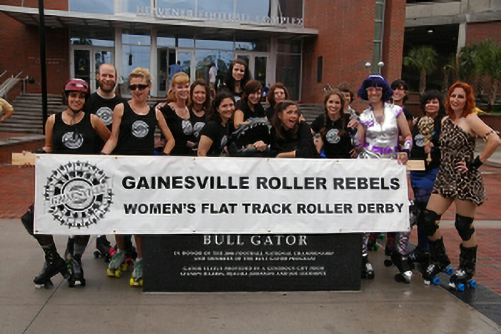 GRR poses with a banner that says "Gainesville Roller Rebels Womens Flat Track Roller Derby" next to the bull gator statue on the University of Florida campus