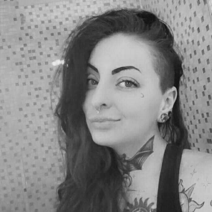 Black and white photo of Nuclear posing in front of polka dotted wallpaper. She has long hair with an undercut and several tattoos on her neck, chest, and arms
